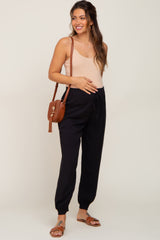 woman wearing a beige maternity tank top and black maternity jogger pants, holding her baby bump and carrying a brown bag