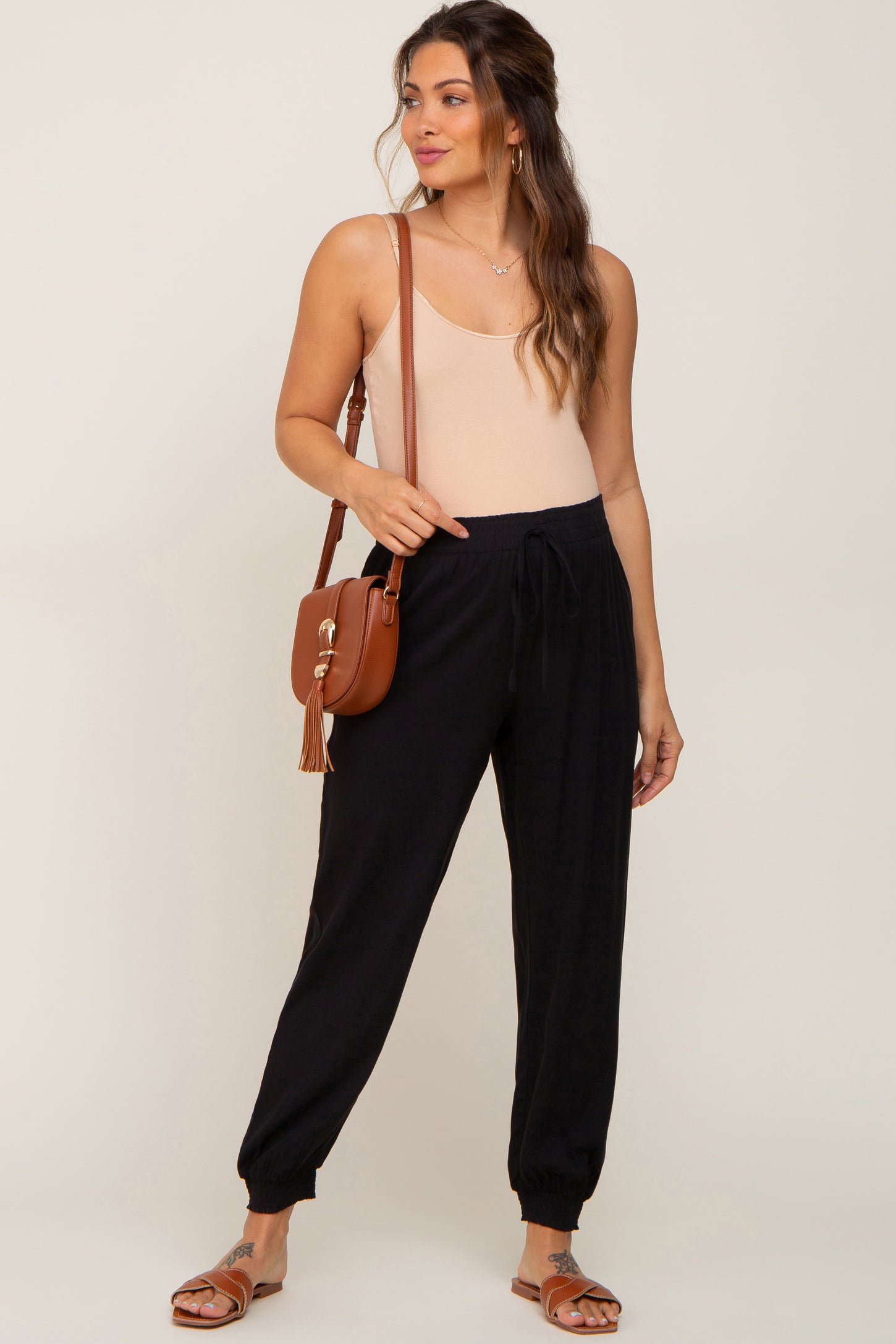 woman wearing black maternity joggers and a light beige tank top, carrying a brown crossbody bag