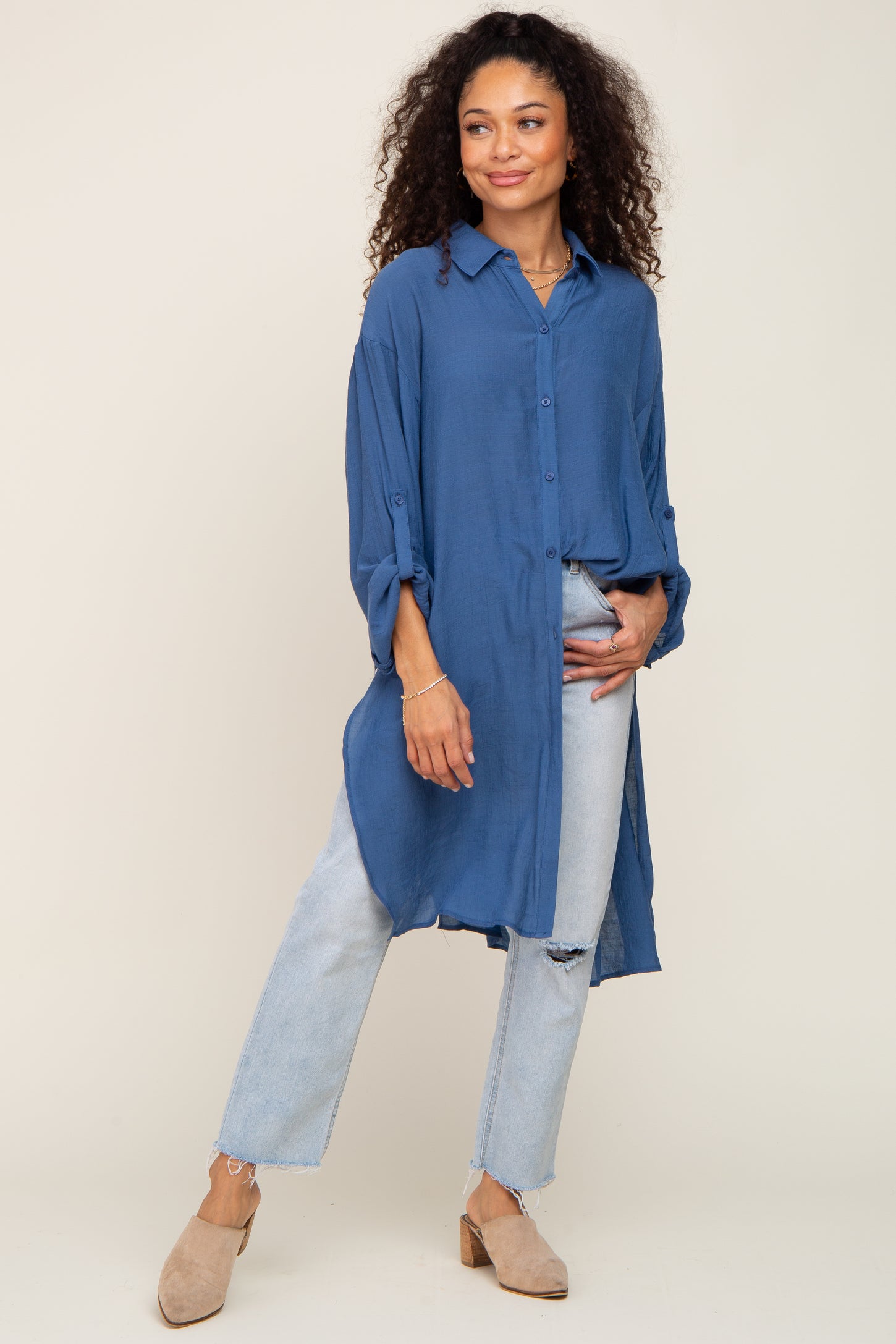 Woman wearing a long blue button-down tunic shirt with light wash distressed jeans and tan mules