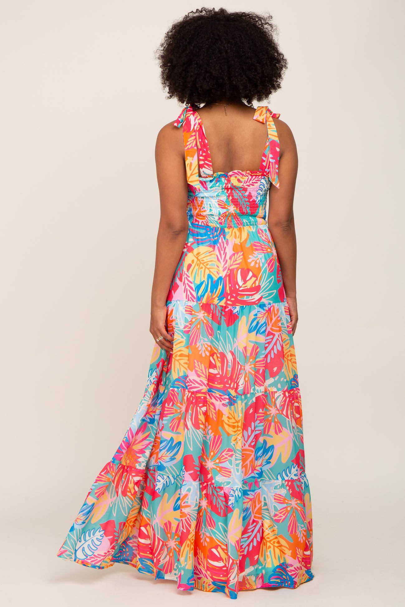 Back view of a woman wearing a colorful tropical floral print tiered maxi dress with tie straps.