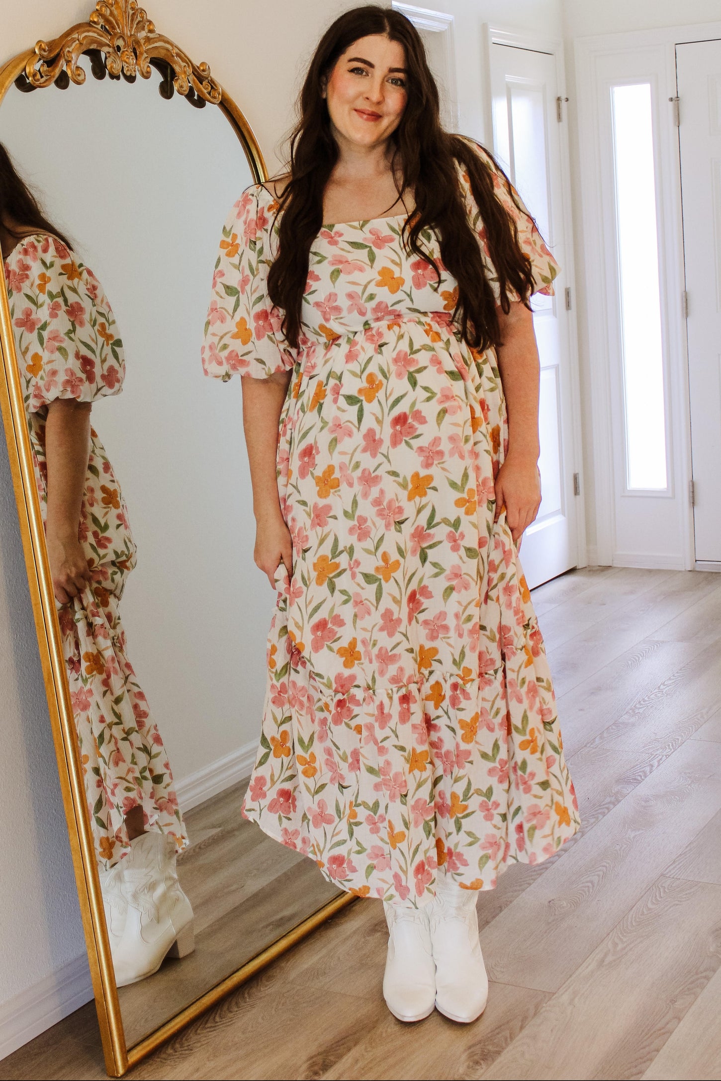 woman wearing a floral maxi dress with puff sleeves and a tiered skirt, standing in front of a mirror with white boots on