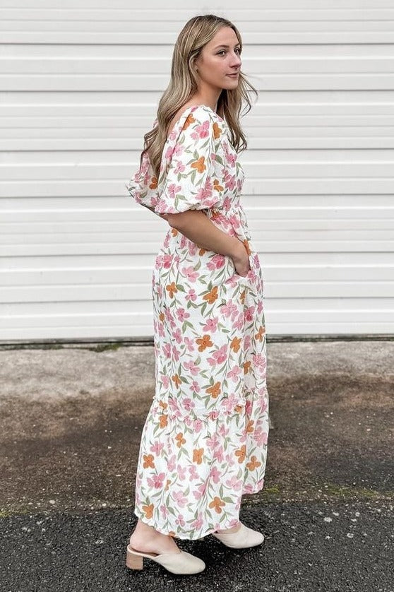 woman wearing a floral maxi dress with puff sleeves and pockets, standing on a pathway near a white backdrop