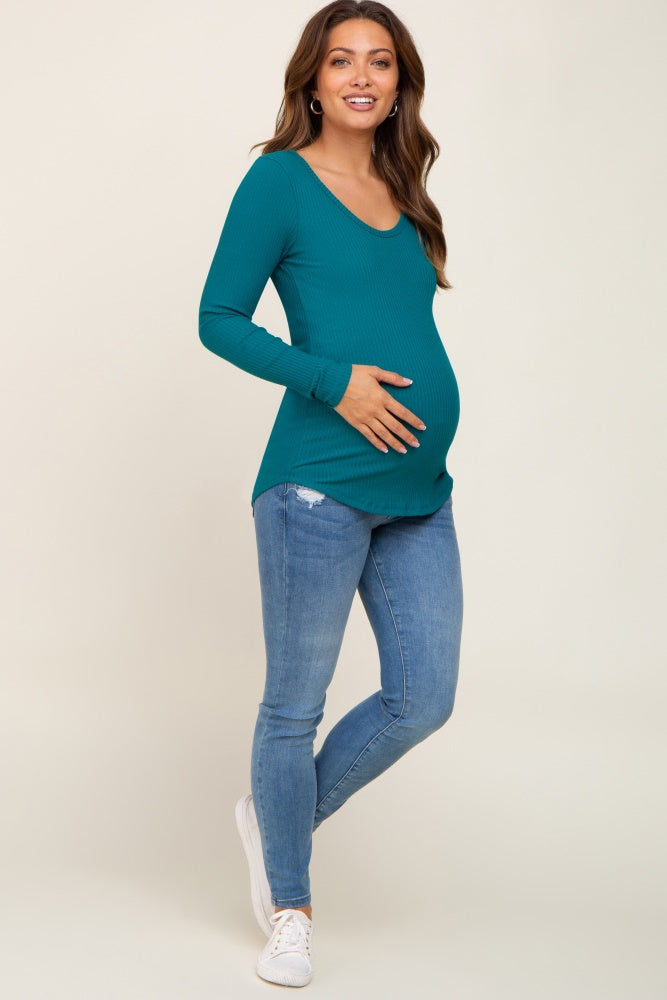 woman wearing a teal ribbed maternity top and blue jeans, cradling her baby bump
