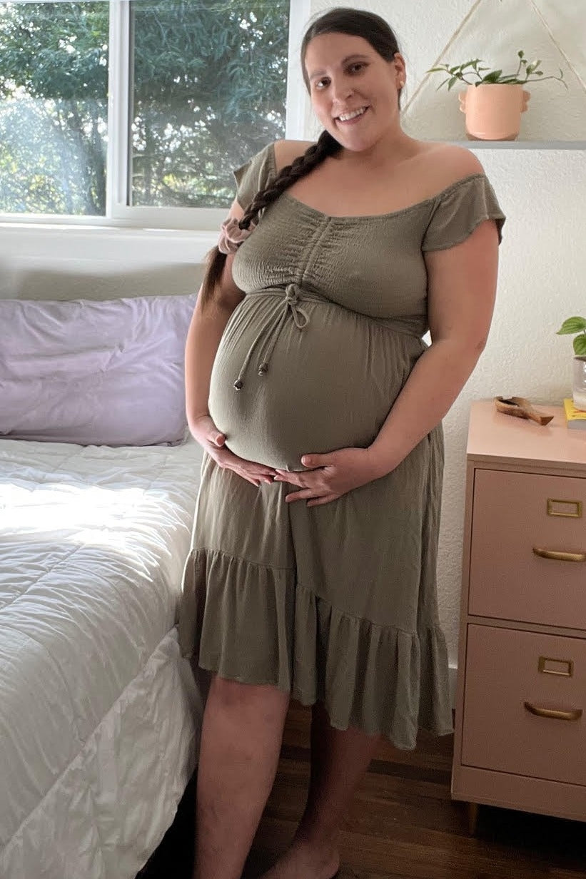 woman wearing an olive green maternity dress with a ruffled hem and smocked bodice, holding her baby bump