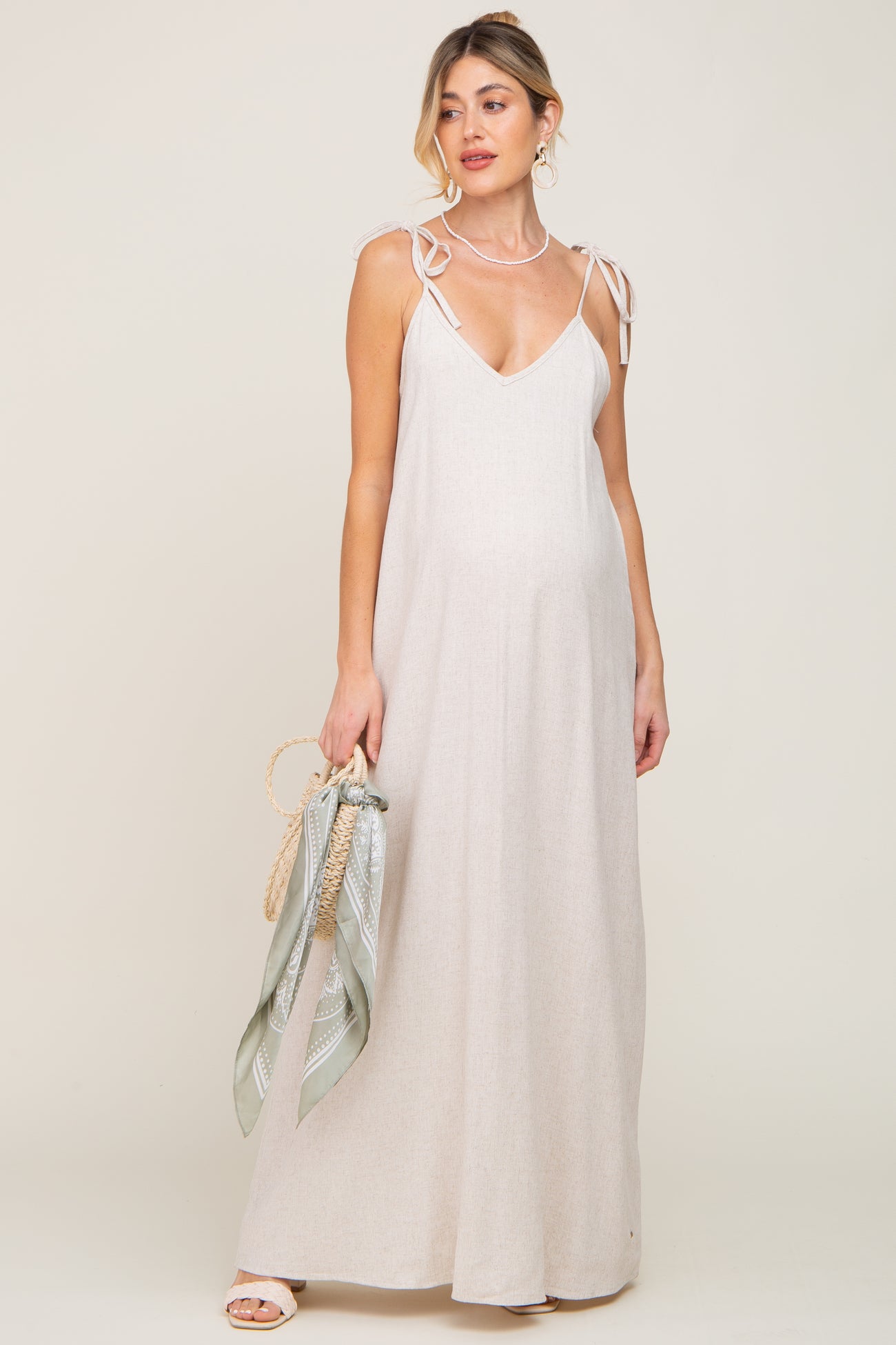 Pregnant woman in a beige linen maternity maxi dress with tie straps holding a straw bag