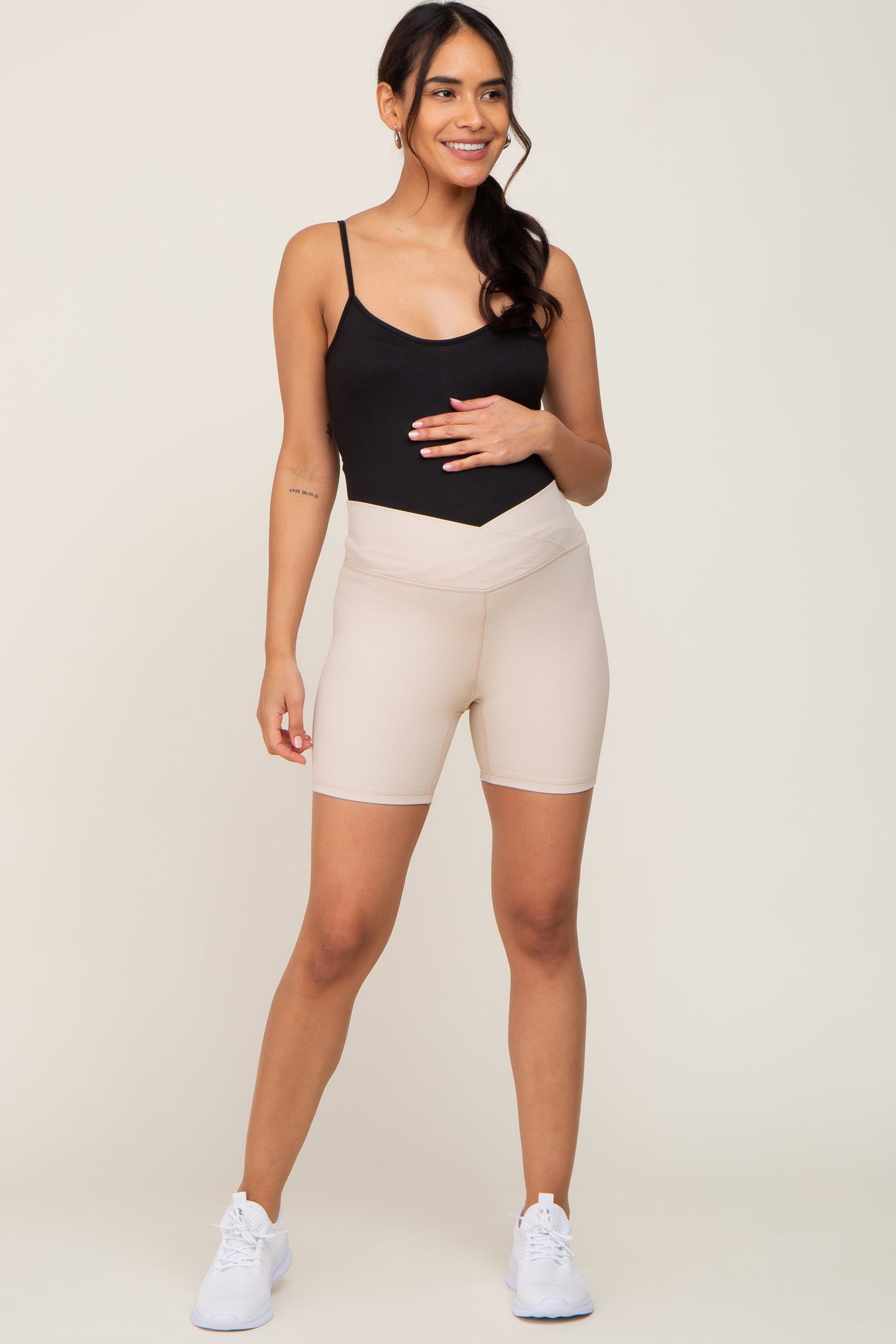 woman wearing a black maternity bodysuit and beige ribbed maternity shorts, holding her baby bump