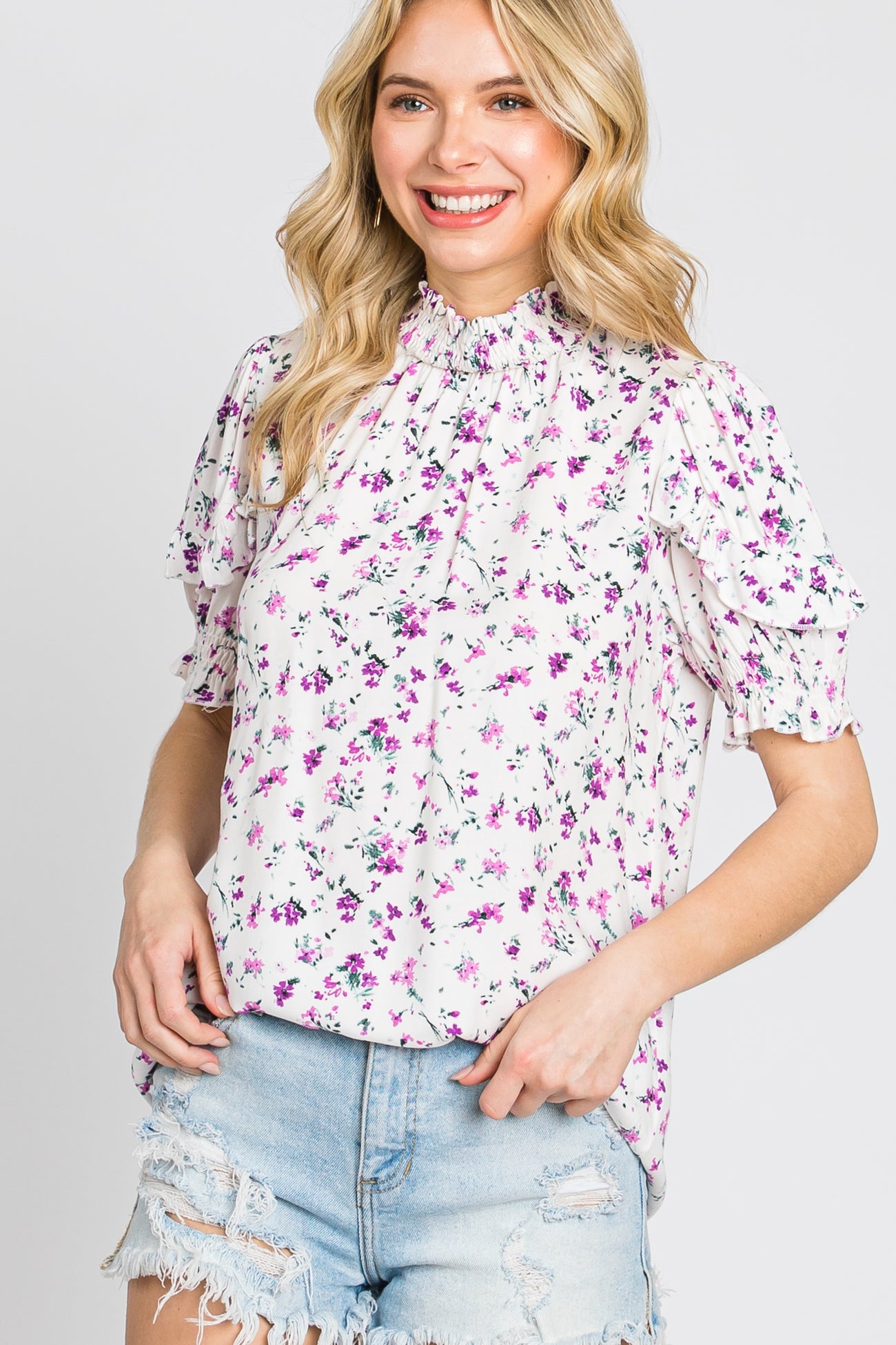 woman wearing a white floral mock neck blouse with purple flowers and distressed denim shorts