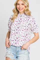 woman wearing a white floral mock neck blouse with purple flowers and distressed denim shorts
