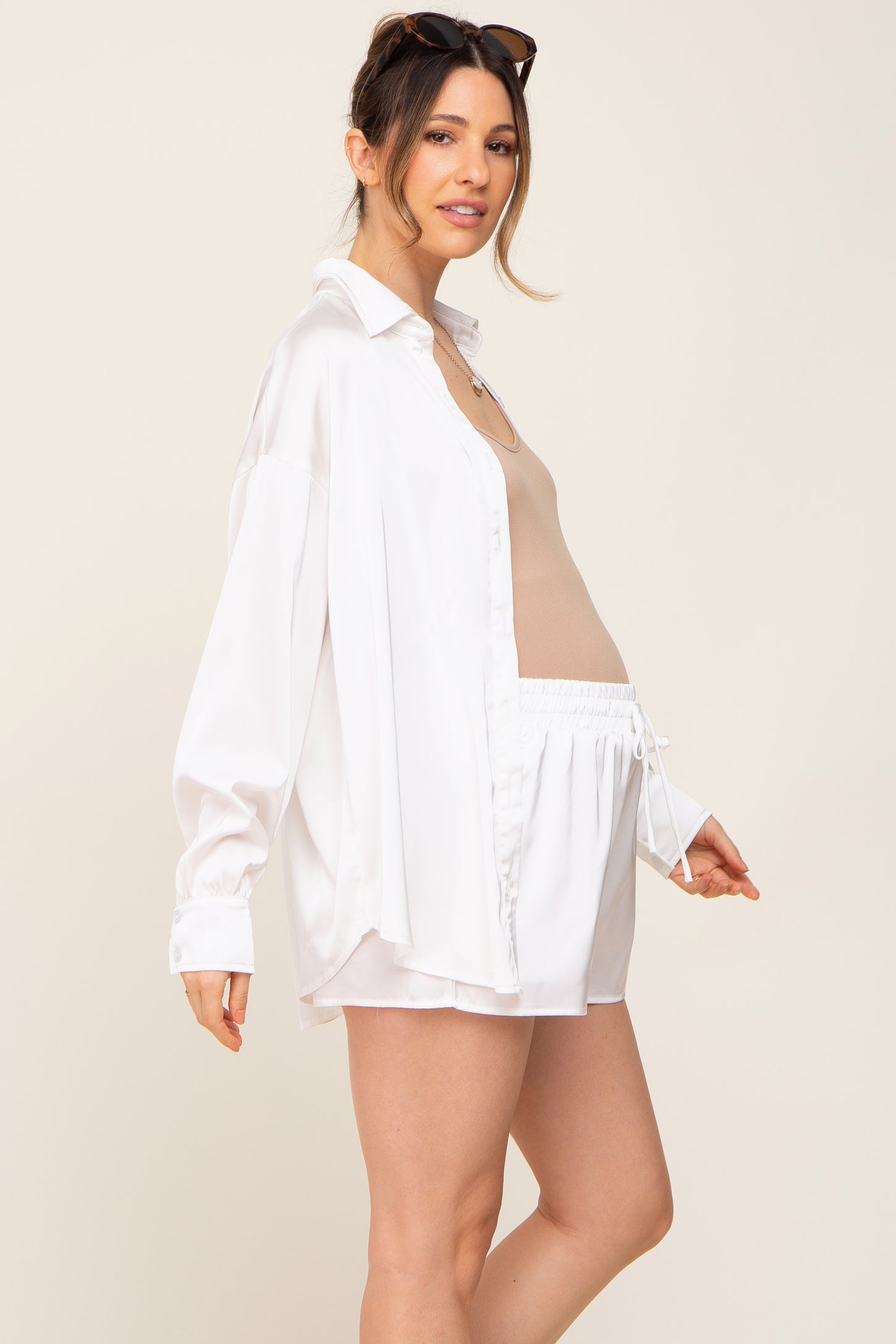 Pregnant woman wearing white satin button-down shirt and matching shorts with a tan maternity tank top