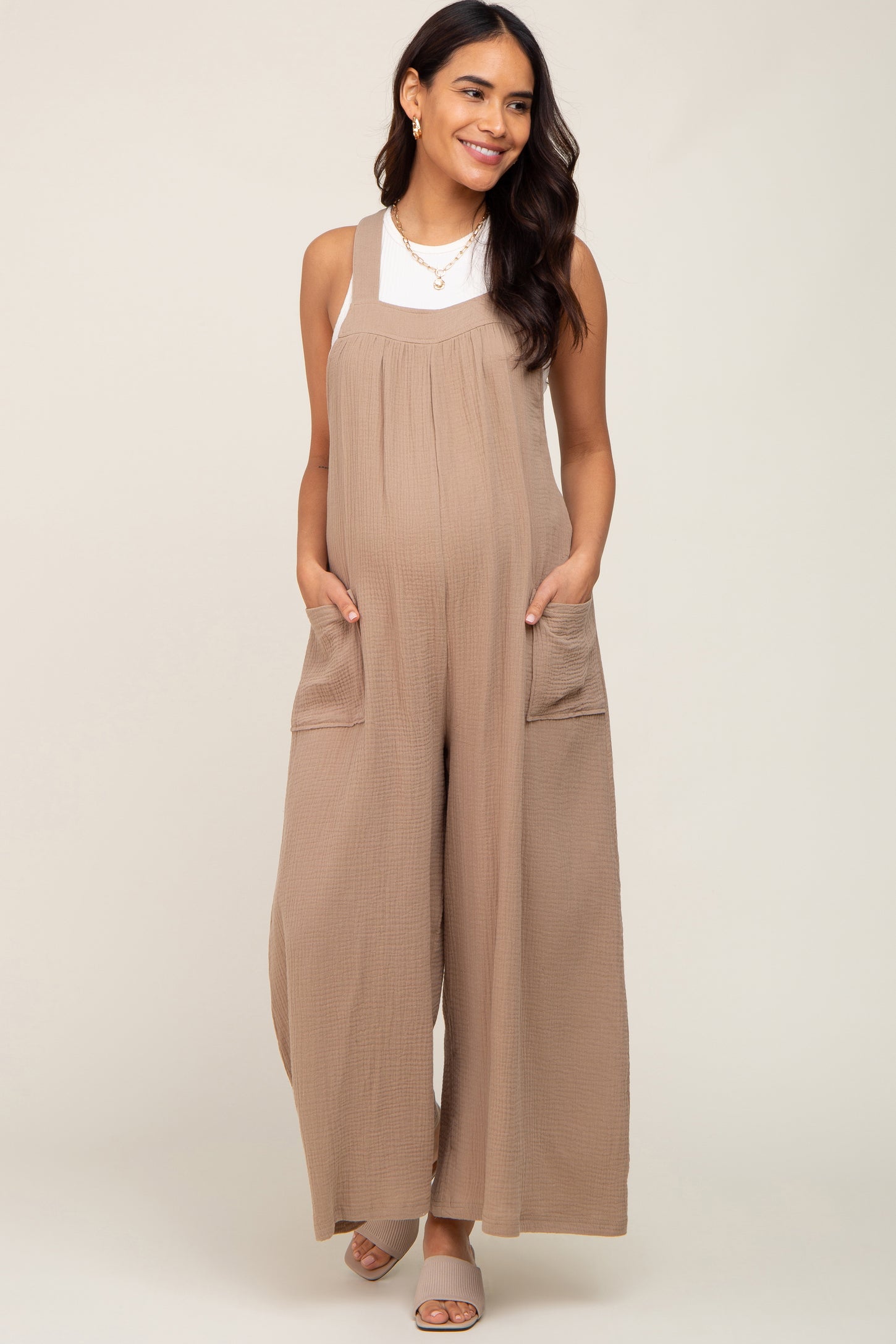 pregnant woman wearing a tan gauze maternity jumpsuit with wide legs and side pockets over a white top