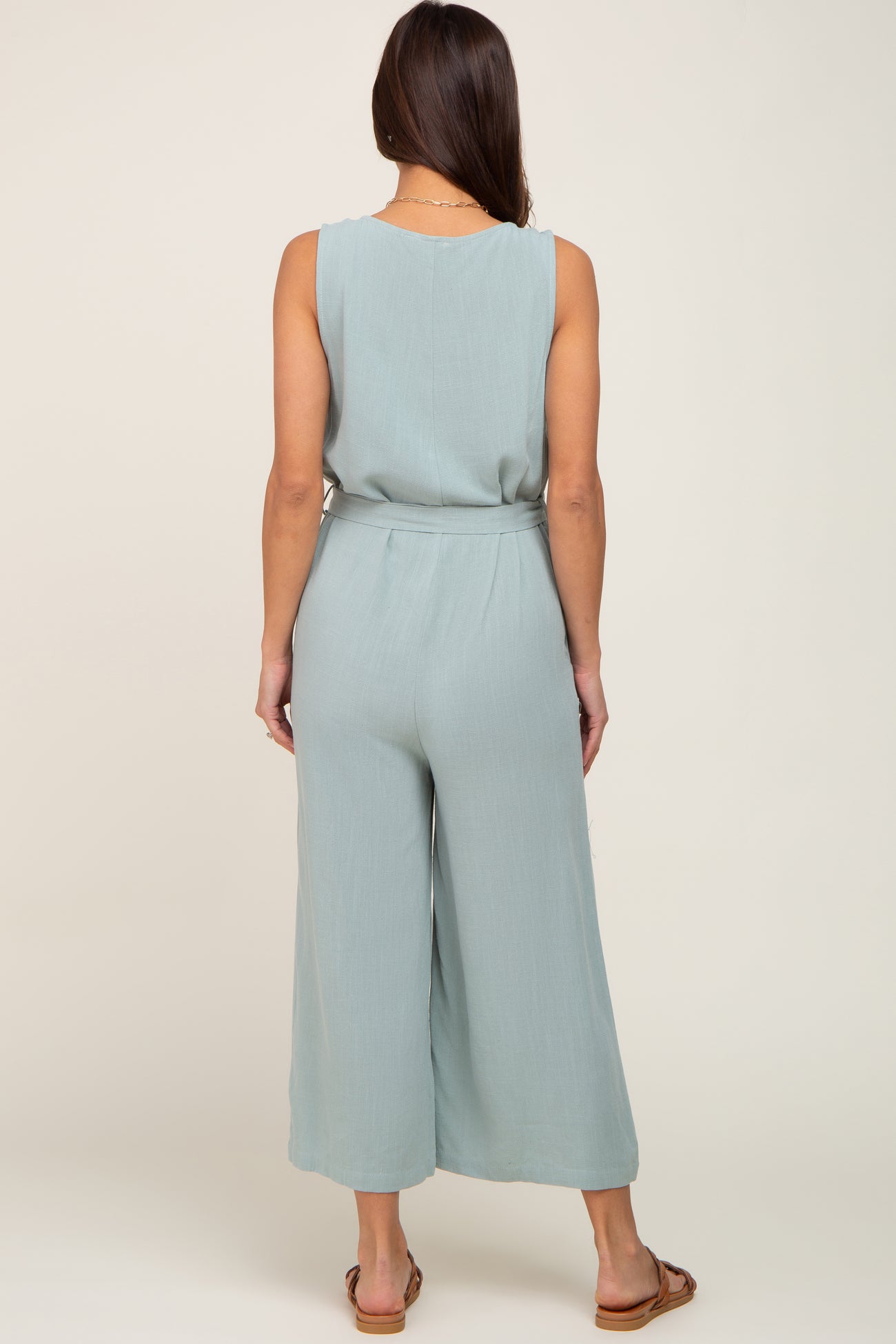 Back view of a woman wearing a light blue sleeveless wide-leg jumpsuit with a belted waist and brown sandals