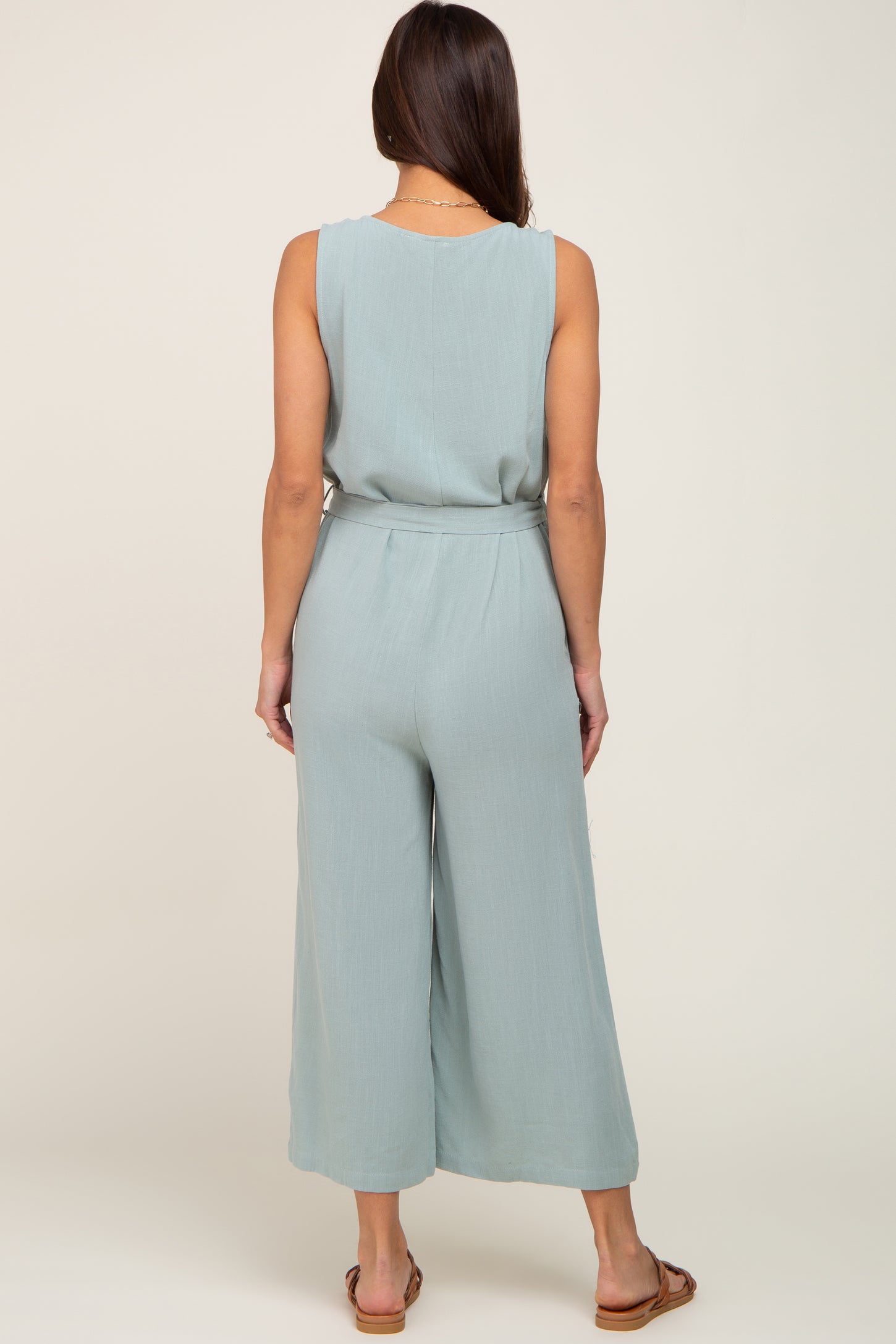 Back view of a woman wearing a light blue sleeveless wide-leg jumpsuit with a belted waist and brown sandals