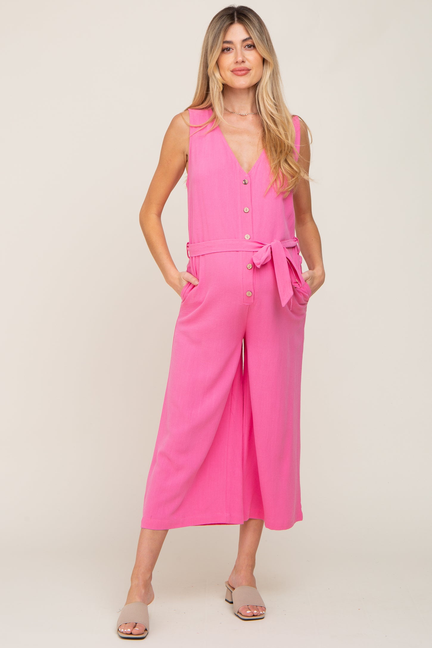 woman wearing a hot pink sleeveless maternity jumpsuit with a belted waist and pockets, showing her baby bump