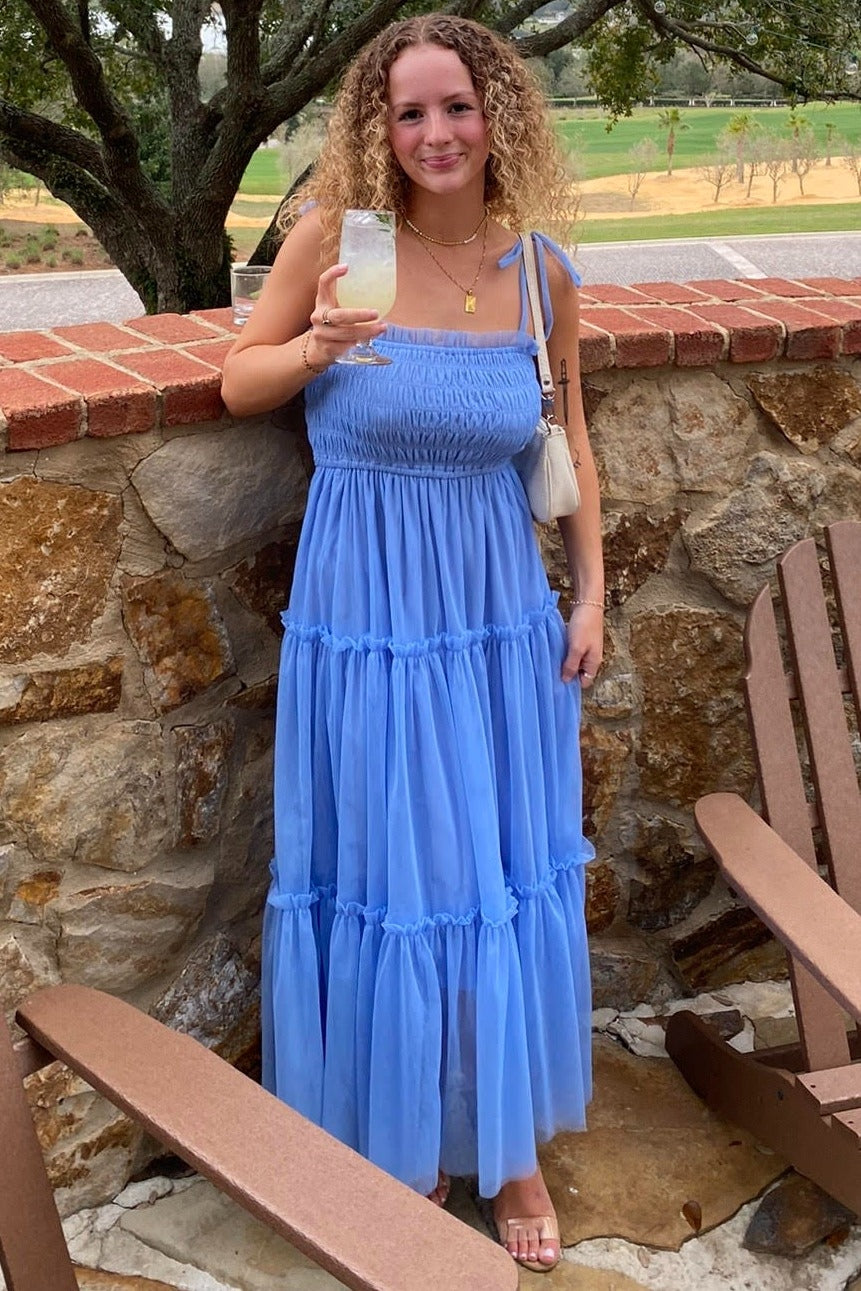 woman wearing a light blue tiered maxi dress with spaghetti straps, holding a drink while standing against a stone wall