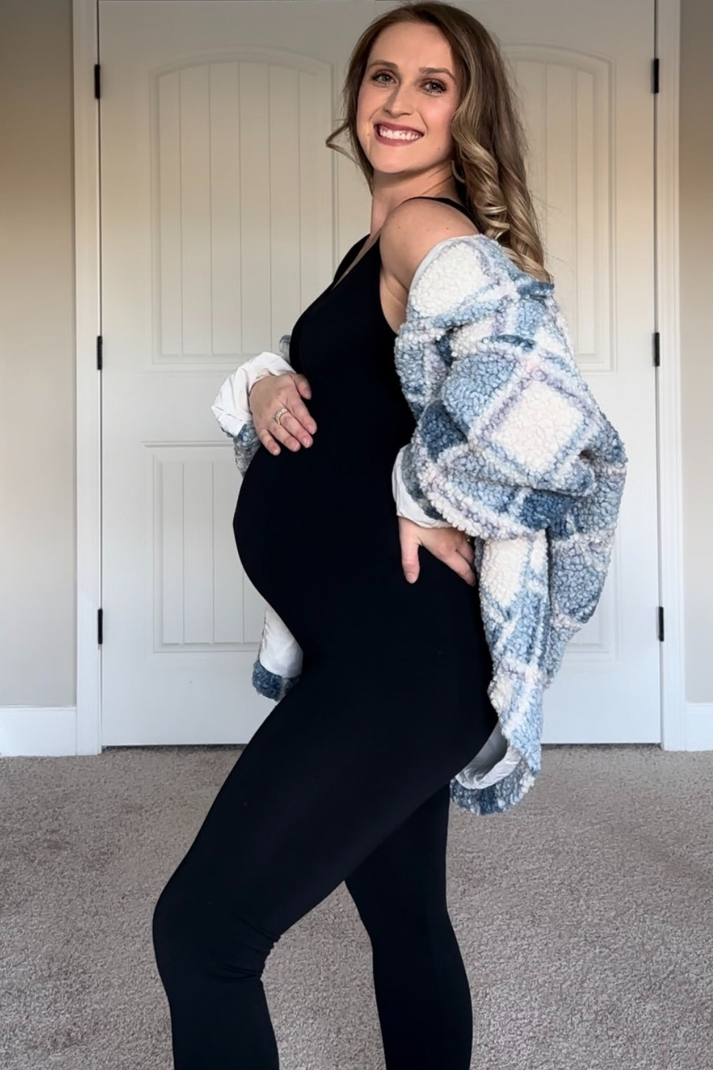 smiling pregnant woman in black maternity jumpsuit and blue plaid sherpa jacket holding her baby bump