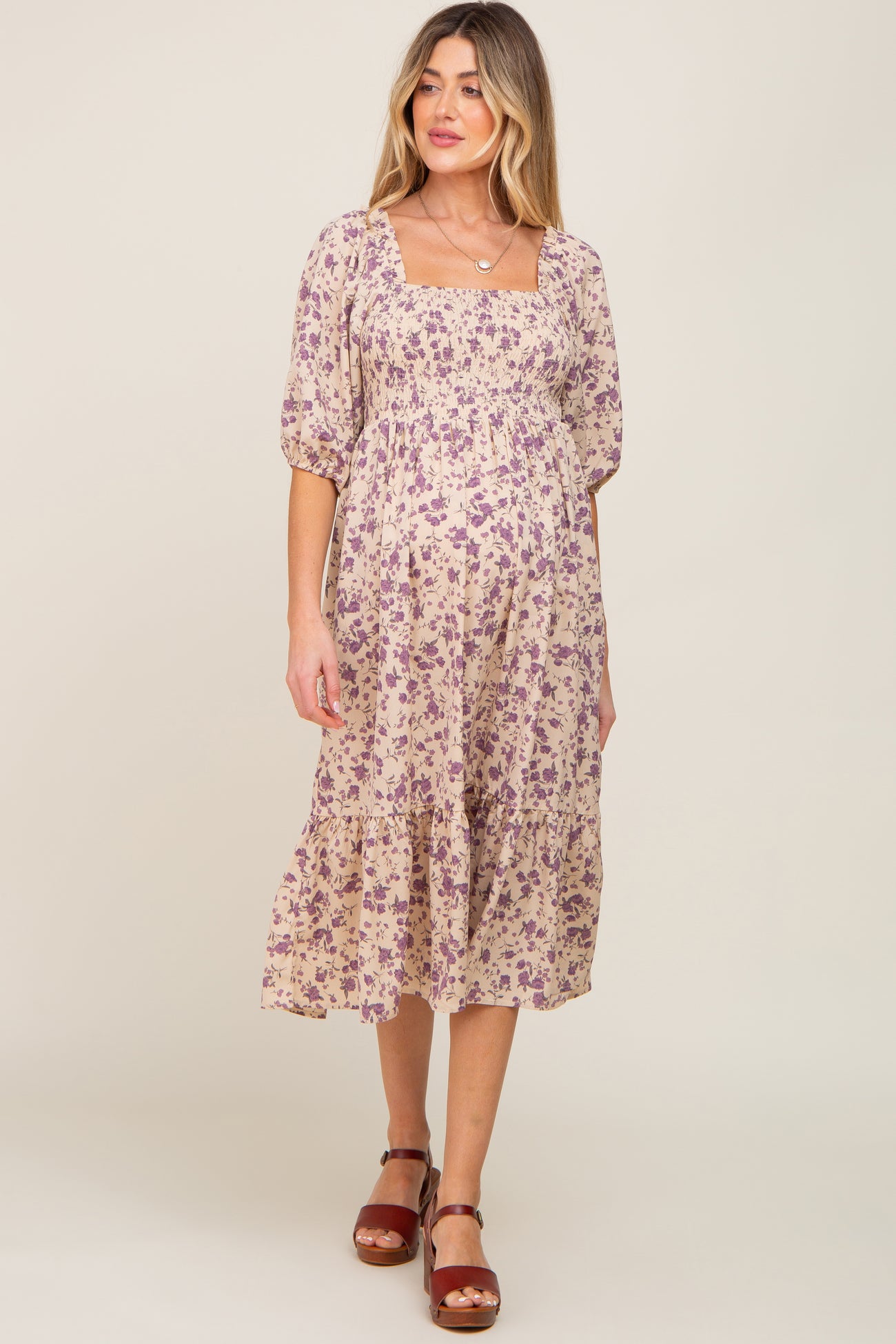 woman wearing a purple floral smocked maternity midi dress with puff sleeves and brown sandals