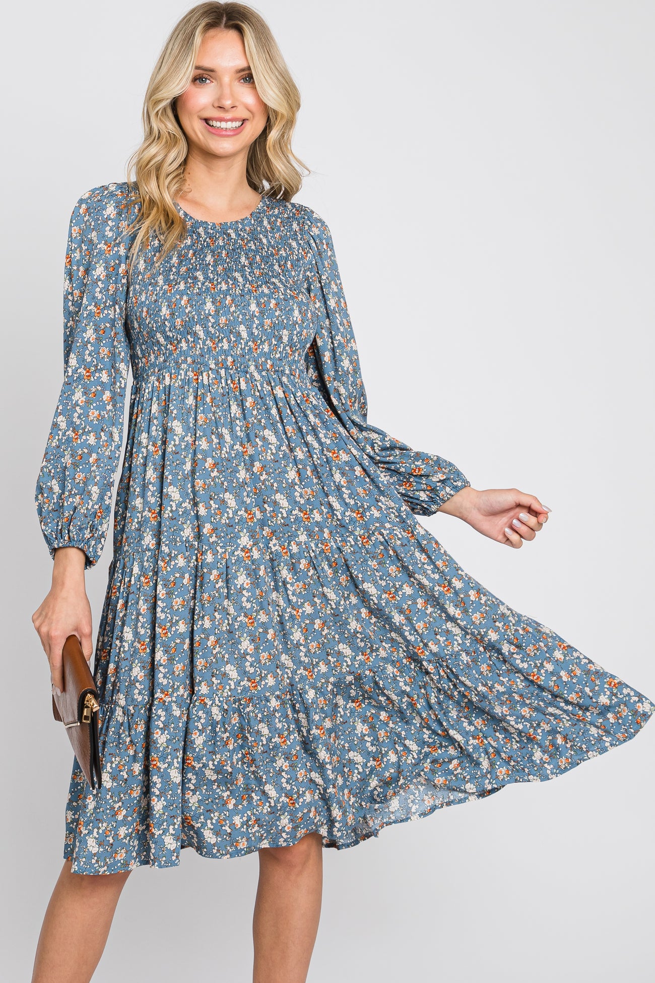 Blonde woman wearing a blue floral smocked midi dress with long sleeves and holding a brown clutch bag