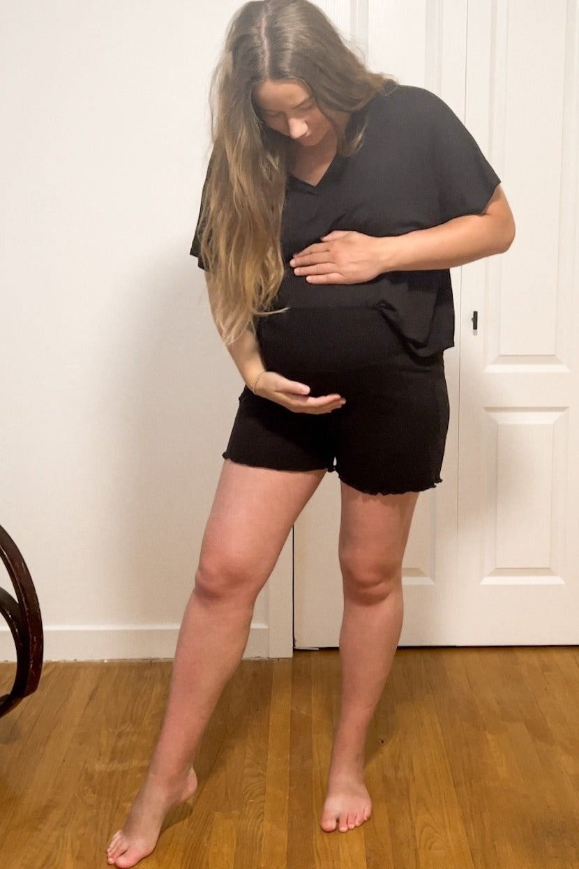 woman wearing a black v-neck maternity top and black shorts, gently cradling her baby bump