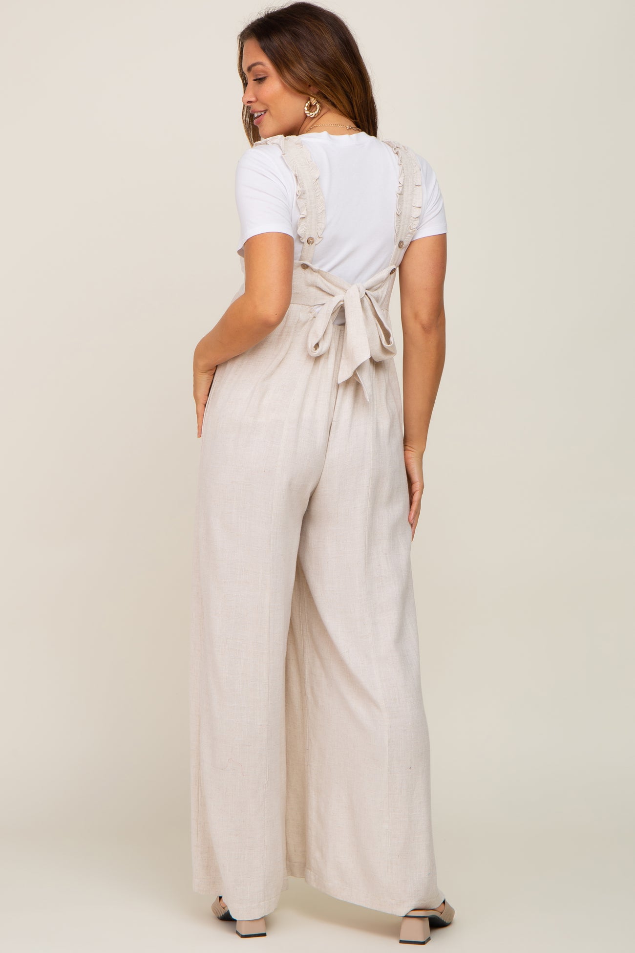 woman wearing beige linen maternity overalls with ruffle straps and back tie over a white t-shirt