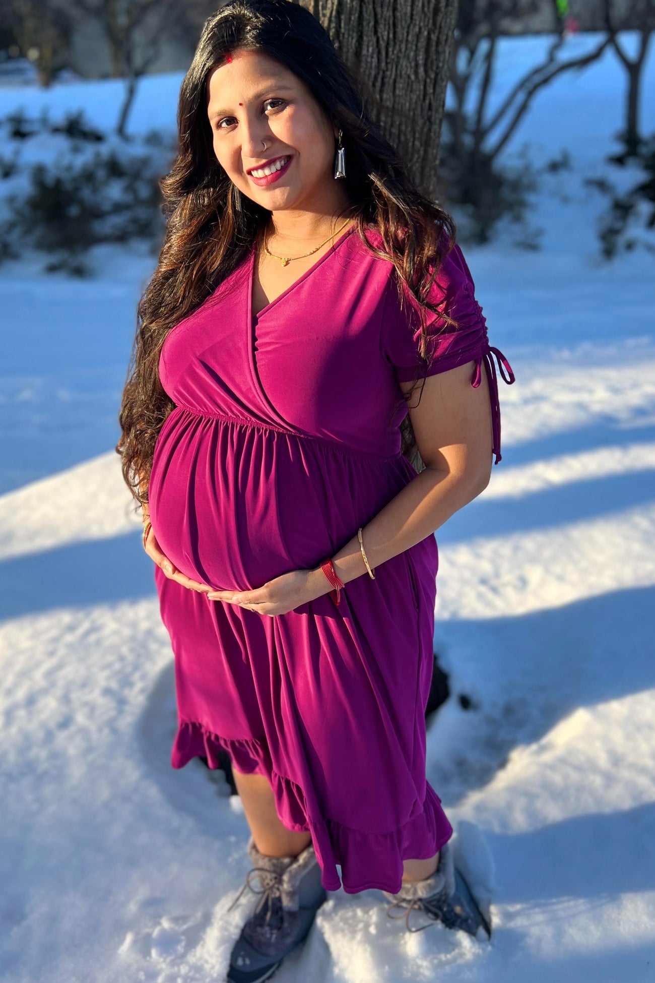 woman wearing a fuchsia short sleeve maternity dress with a ruffled hem, holding her baby bump in a snowy setting