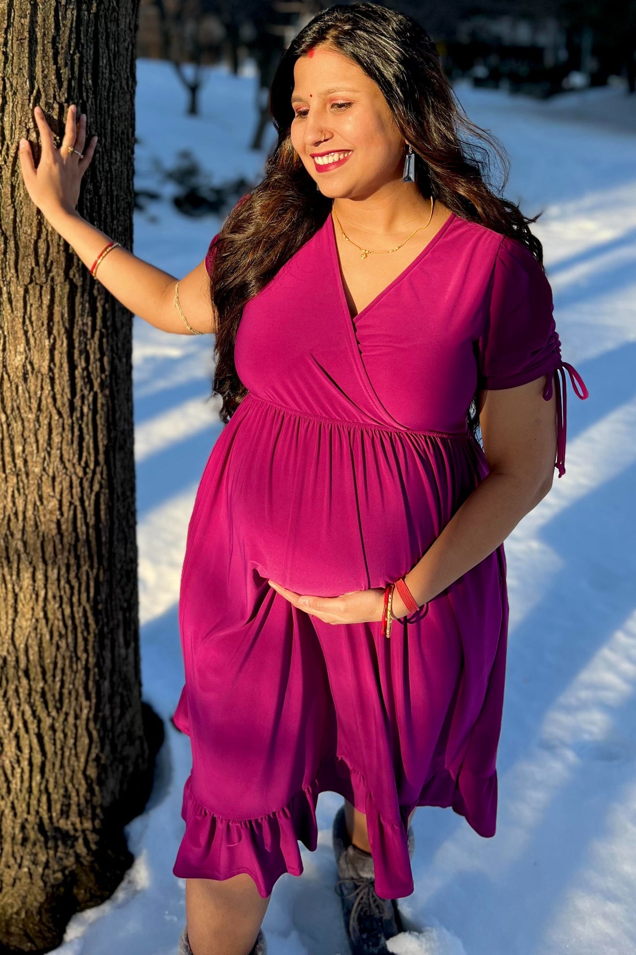 woman wearing a raspberry pink maternity dress with ruffle hem and holding her baby bump in the snow