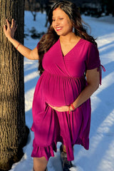 woman wearing a raspberry pink maternity dress with ruffle hem and holding her baby bump in the snow