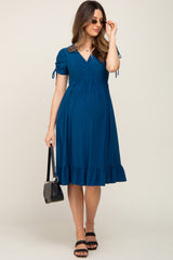 woman wearing a dark teal maternity dress with short sleeves and a ruffled hem, holding a small handbag and wearing sunglasses