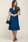 woman wearing a teal maternity dress with short sleeves, cinched waist, and ruffle hem, holding a black handbag