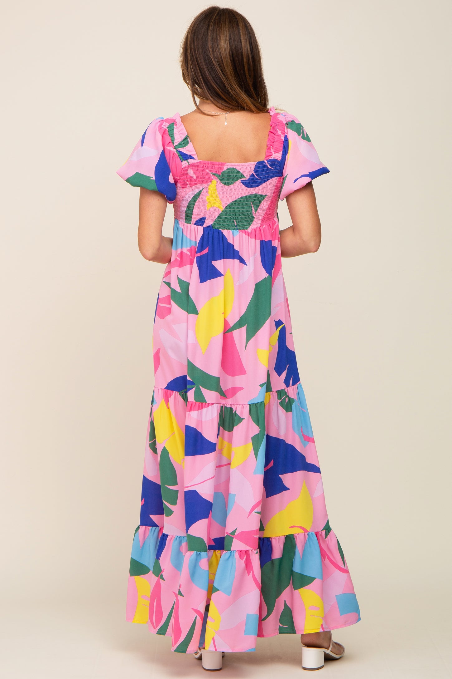 Back view of a woman wearing a pink tropical print tiered maxi dress with smocked bodice and flutter sleeves