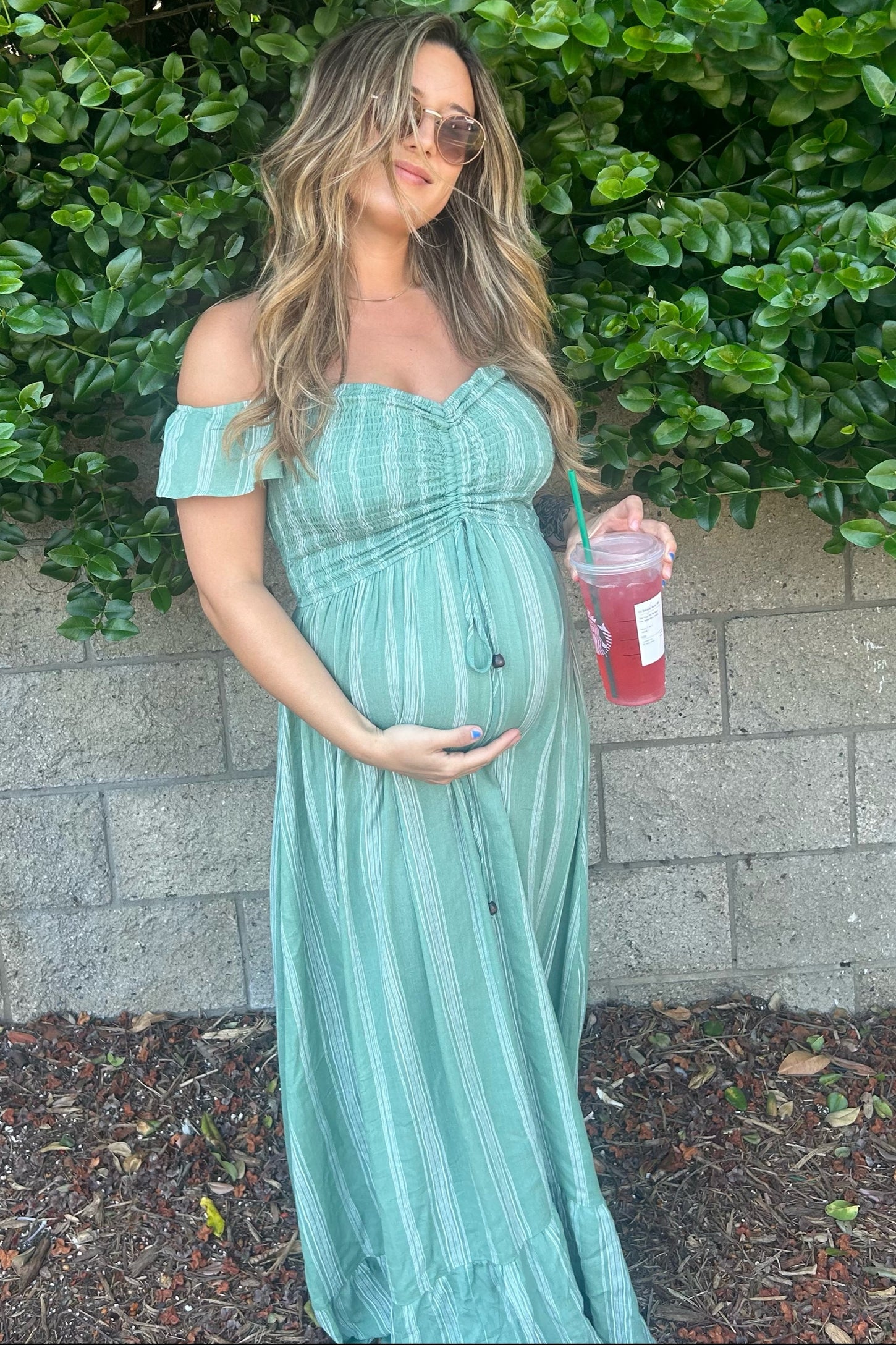 woman wearing a green striped off-the-shoulder maternity maxi dress and holding a drink with her baby bump visible