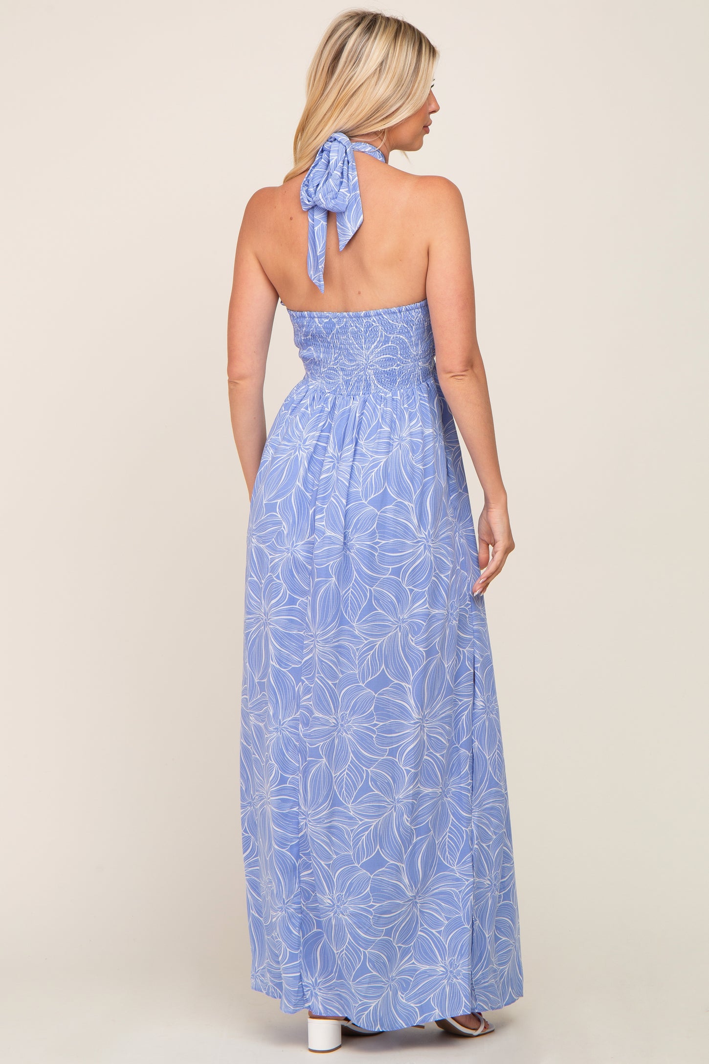 Back view of a woman wearing a light blue floral halter neck maxi dress with a smocked waist and tie neck.