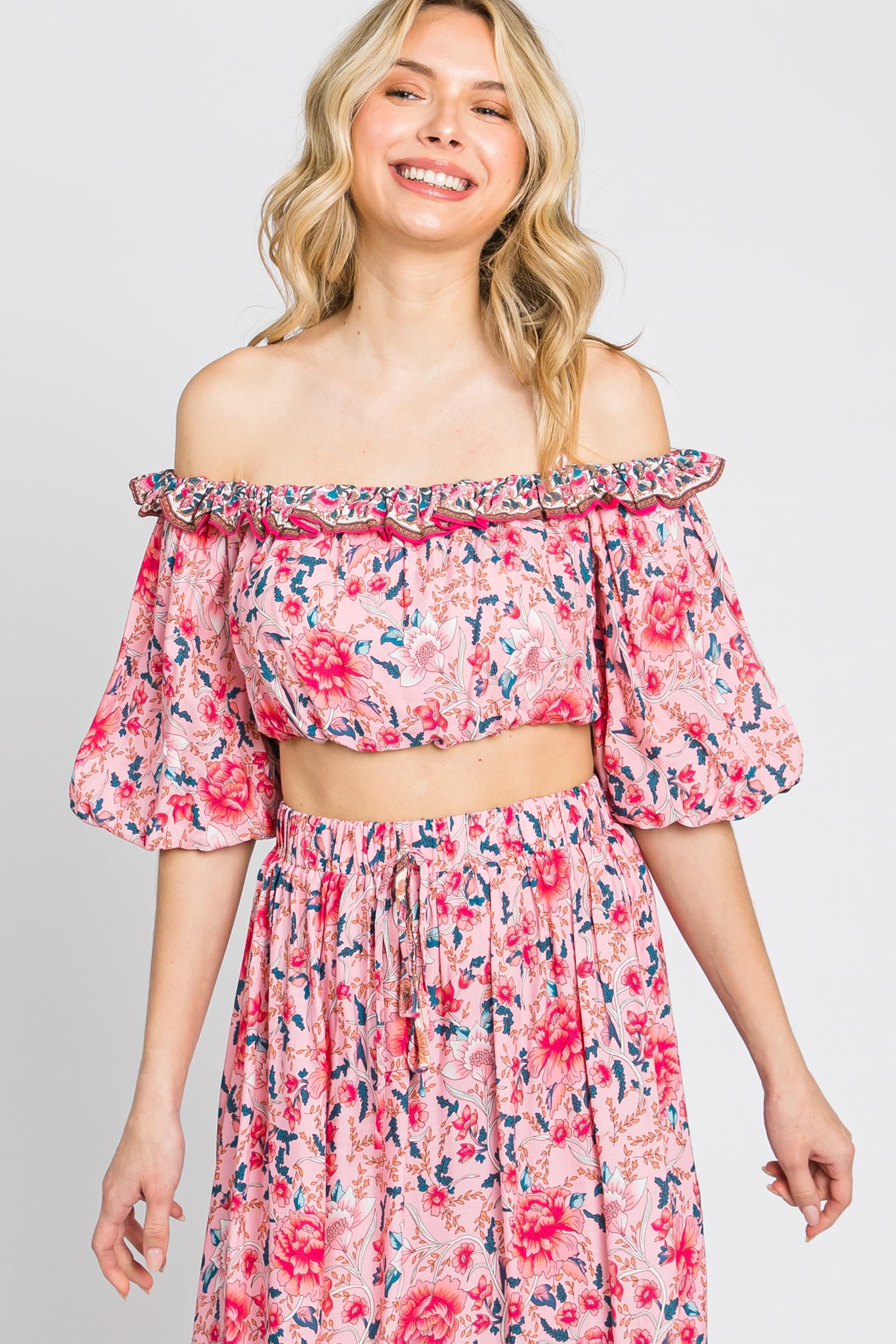woman wearing a pink floral off-the-shoulder crop top with puff sleeves and a matching skirt, smiling at the camera