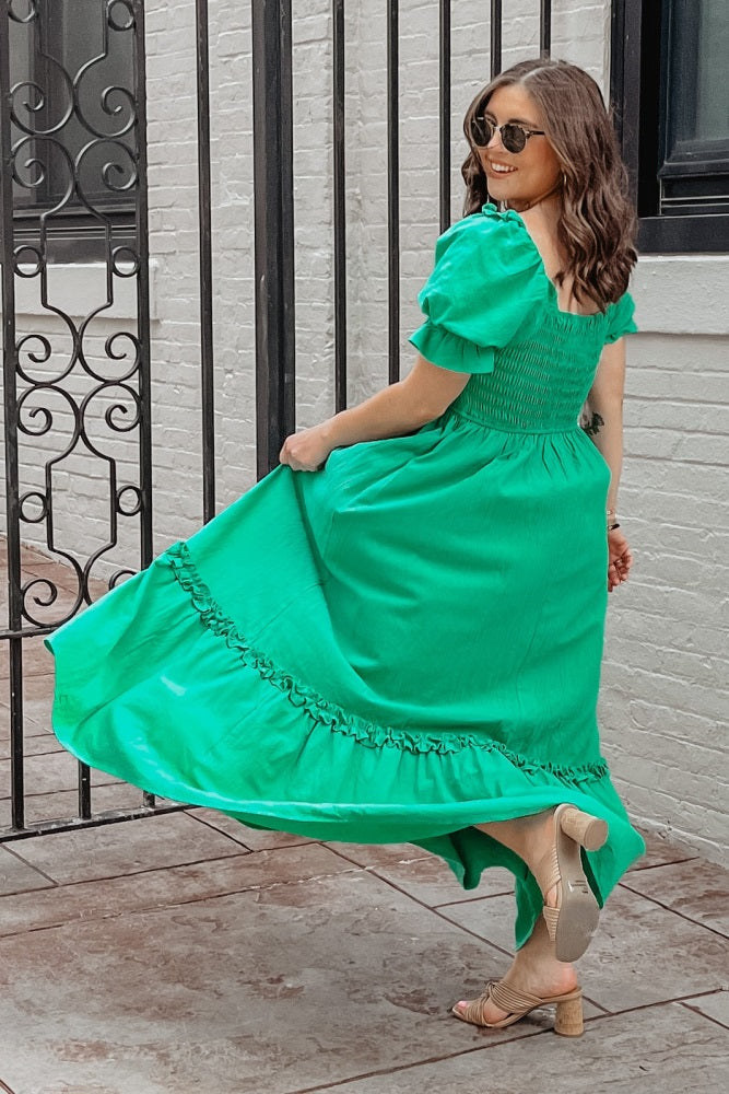 woman in green puff sleeve maxi dress and tan block heel sandals posing by a black wrought iron gate