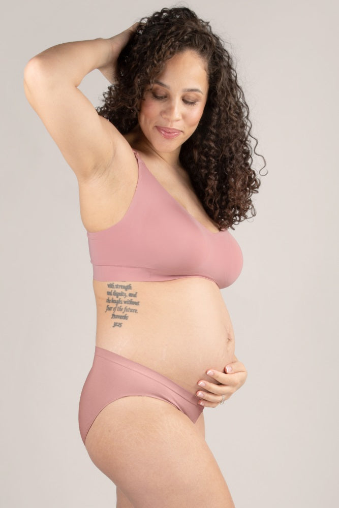 Pregnant woman with curly hair wearing mauve maternity bra and underwear set holding her baby bump