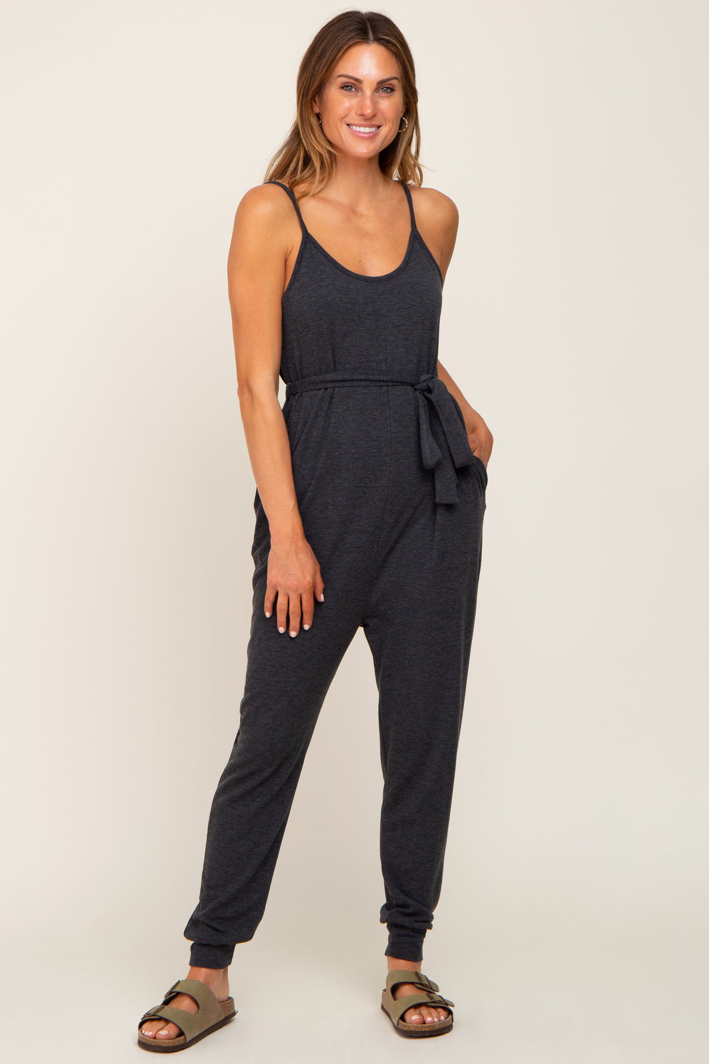 woman wearing a dark gray sleeveless jumpsuit with a tie waist and tapered legs, styled with sandals