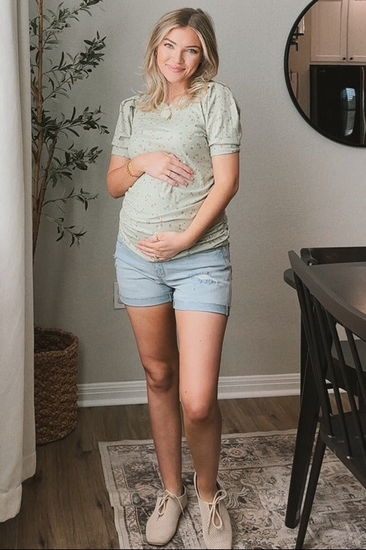 woman wearing a light green maternity top with floral print and distressed denim shorts, holding her baby bump