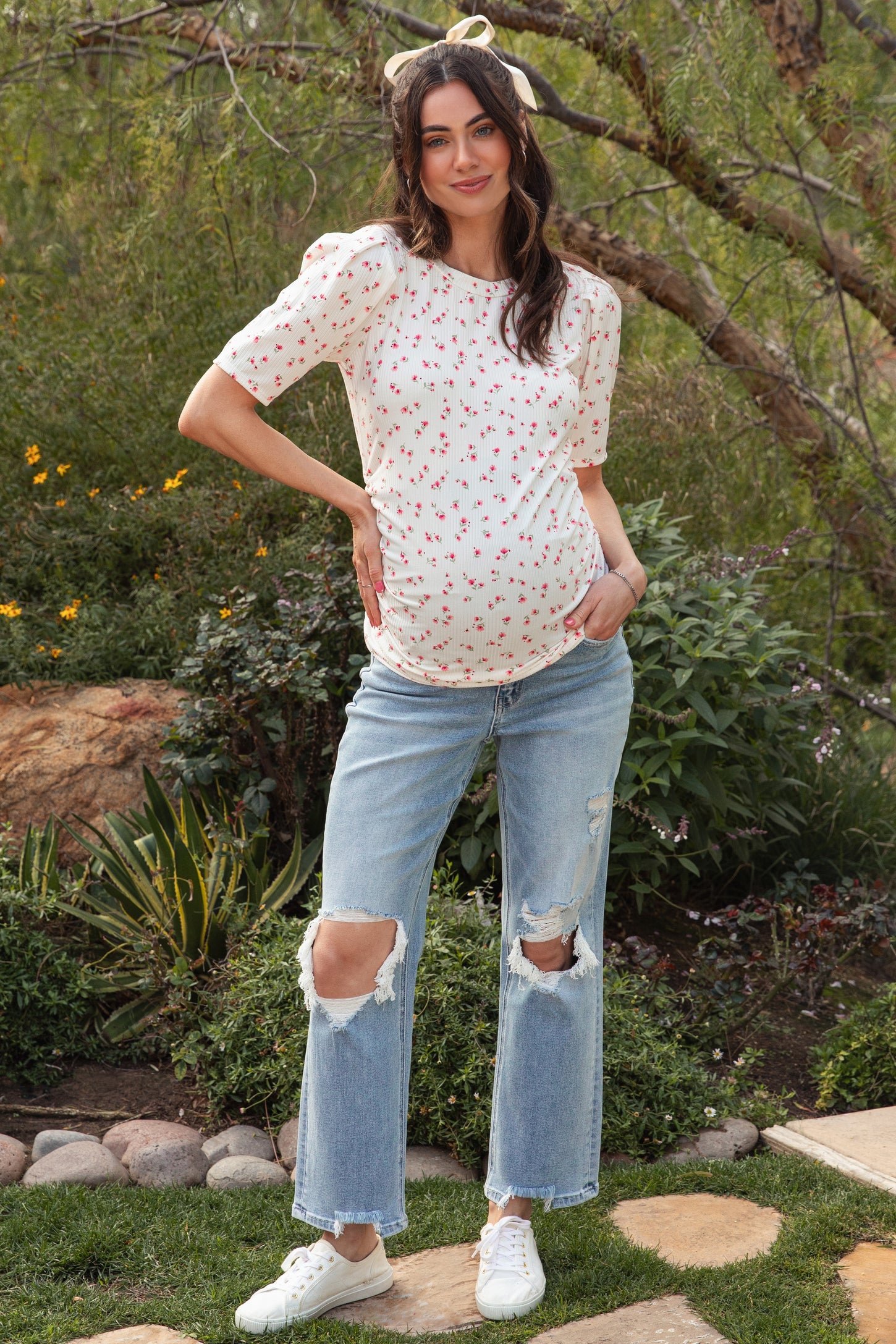 woman wearing a white short sleeve maternity top with red floral print and holding her baby bump, paired with ripped denim jeans