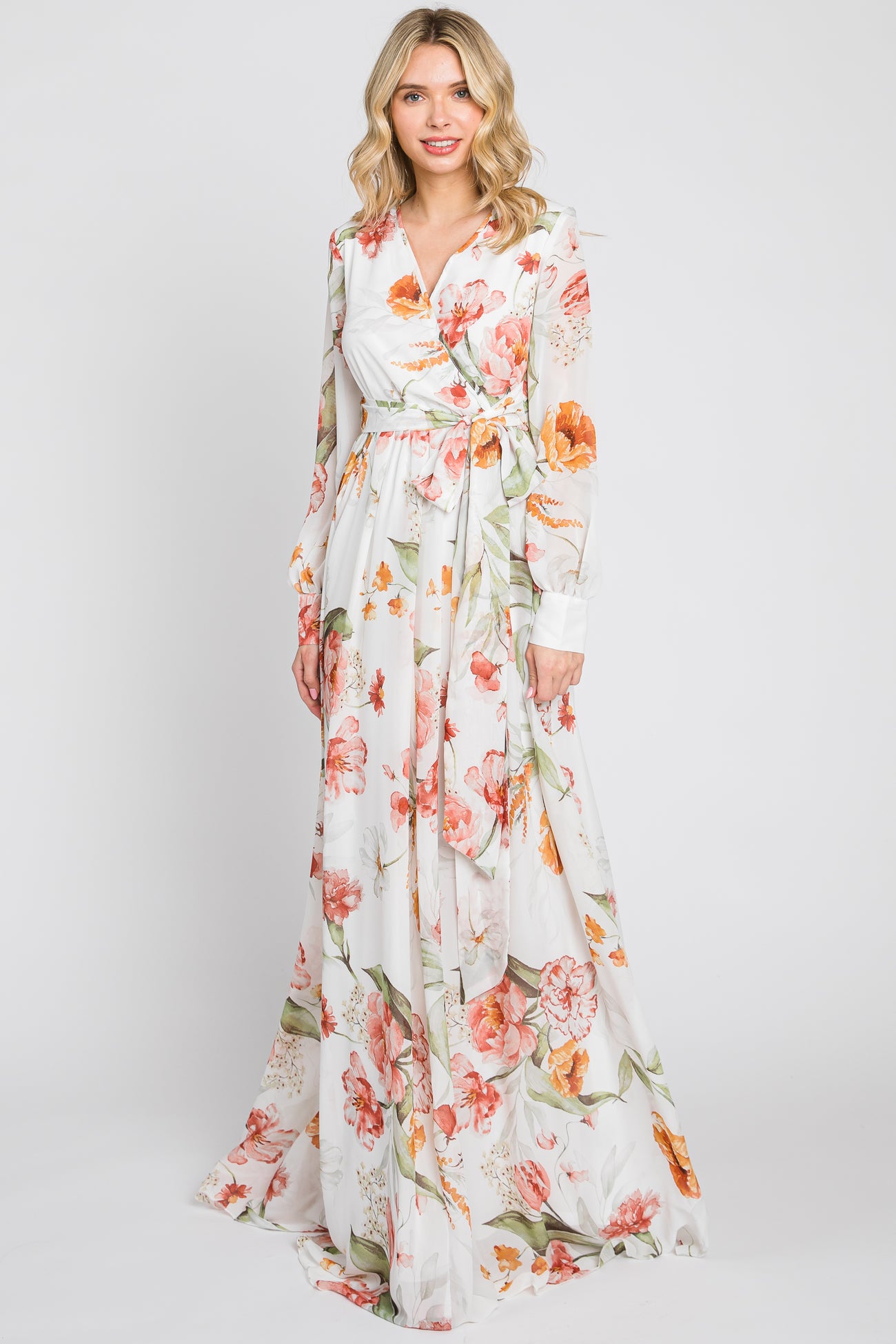Woman wearing a long sleeve white floral maxi dress with orange and pink flowers and a waist tie