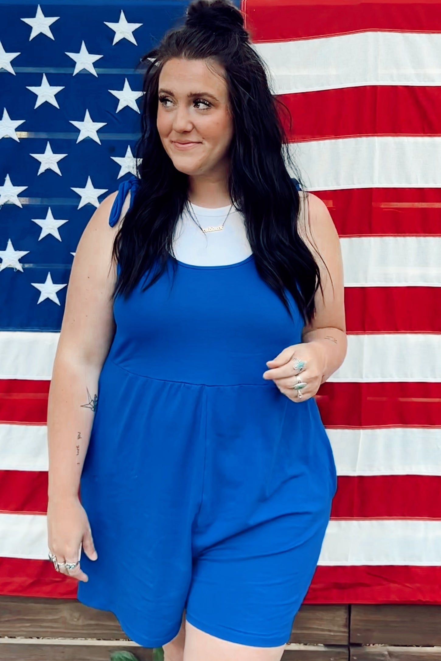 woman wearing a bright blue sleeveless romper with side tie details, standing in front of an American flag backdrop
