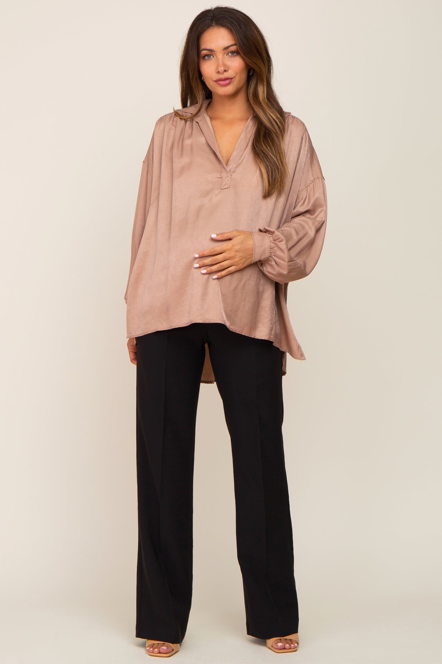 woman wearing a beige maternity blouse with a loose fit and long sleeves, cradling her baby bump with black pants