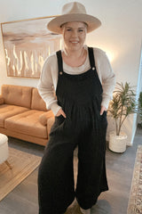 Woman wearing black wide-leg overalls, a cream sweater, and a tan wide-brim hat in a modern living room