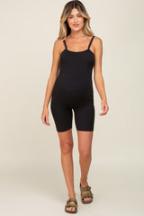 Pregnant woman wearing a black ribbed maternity romper and olive green sandals against a neutral background