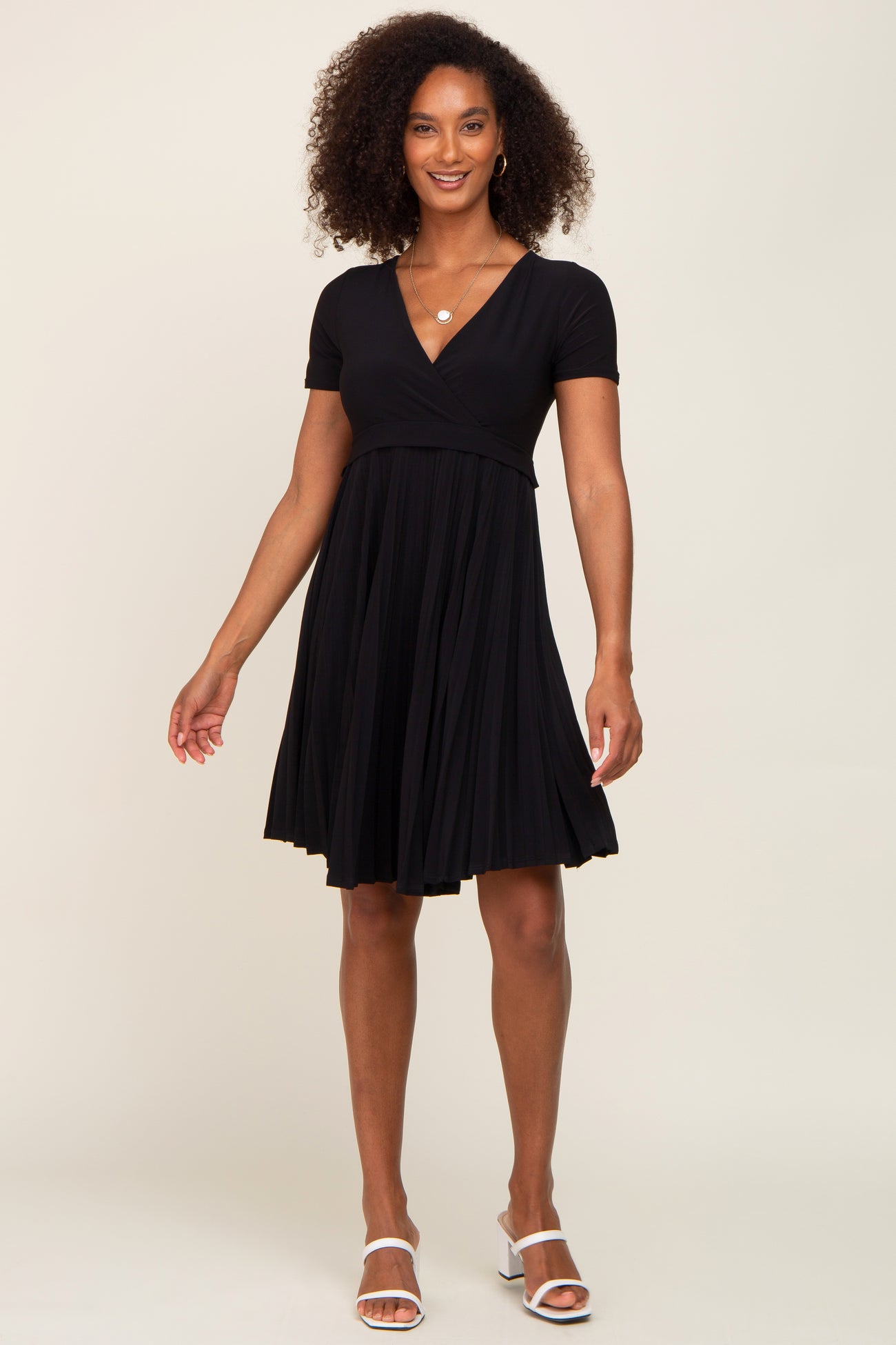 woman wearing a short sleeve black pleated maternity dress with a v-neckline and white block heel sandals