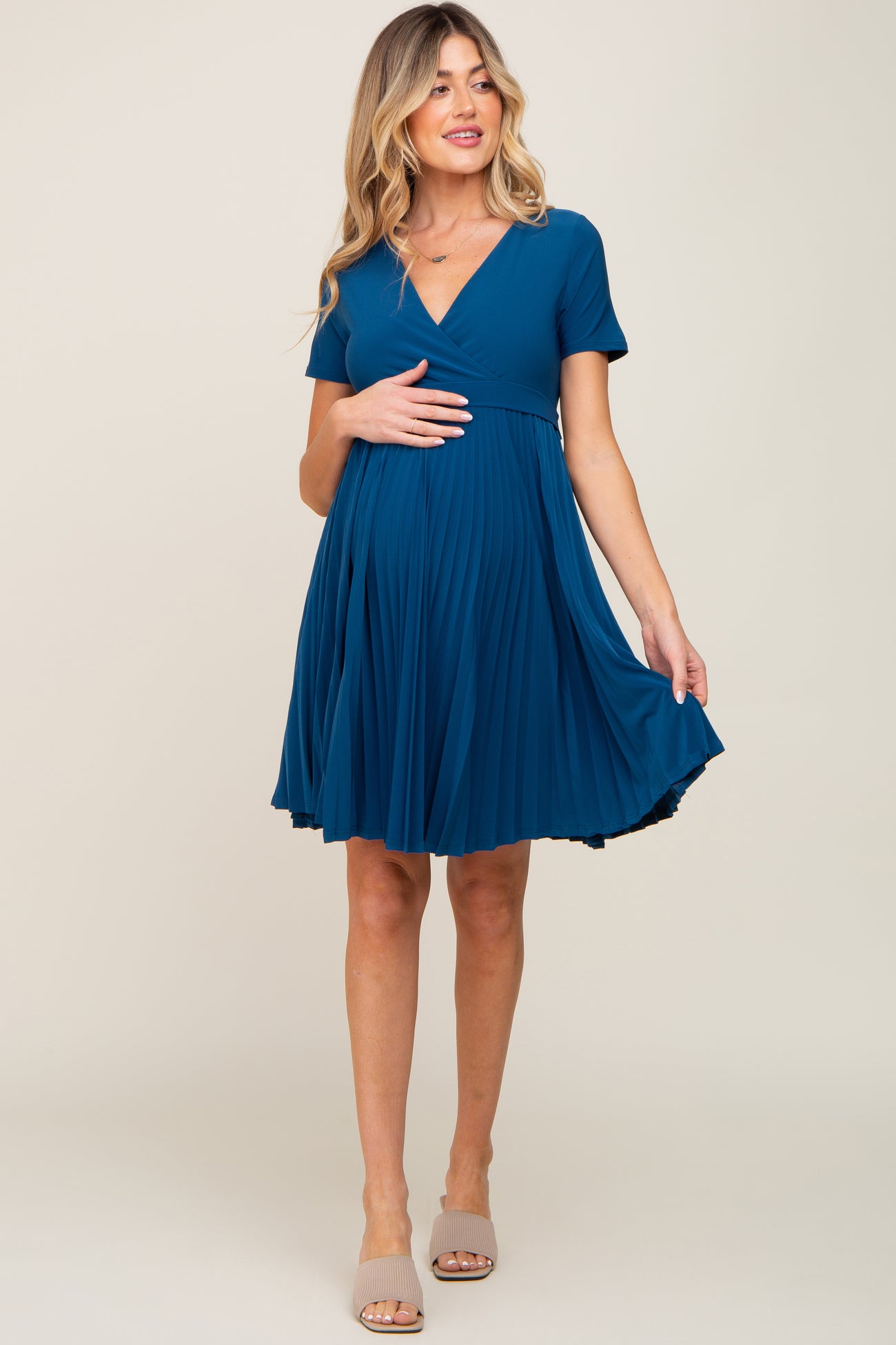 woman wearing a teal maternity dress with short sleeves and a pleated skirt, holding her baby bump