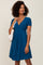 woman wearing a teal pleated wrap dress with short sleeves and a v-neck, holding a white handbag