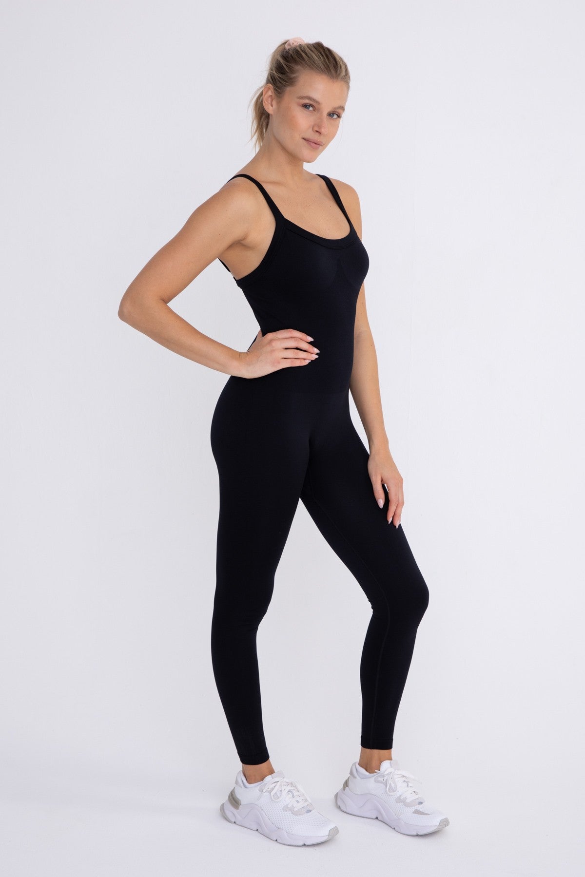 woman wearing a black sleeveless one-piece yoga bodysuit and white sneakers against a white background