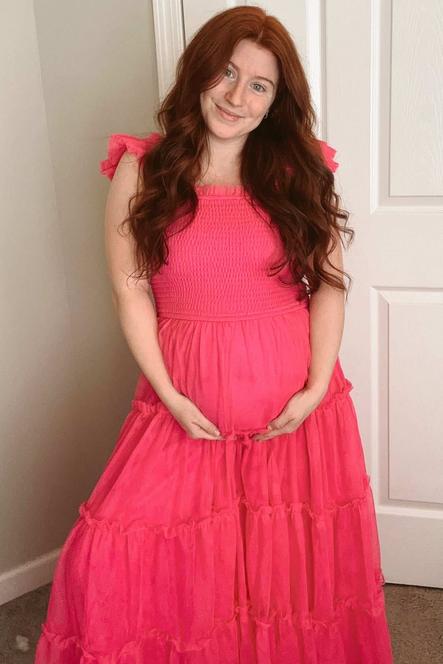 woman wearing a bright pink maternity dress with smocked bodice and ruffled sleeves, holding her baby bump