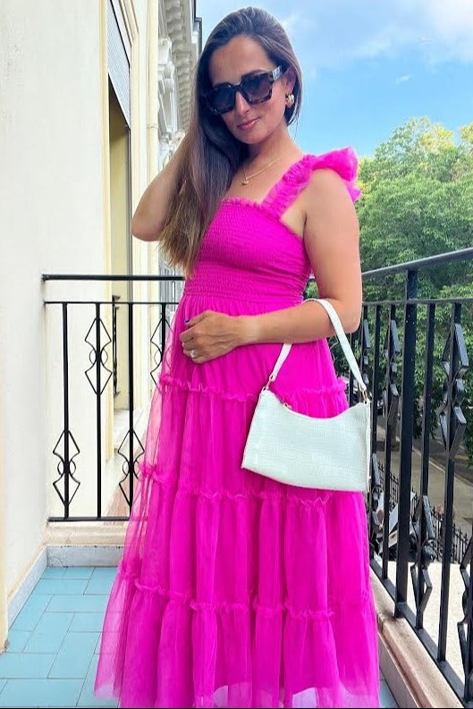woman wearing a hot pink maternity maxi dress with ruffles and sheer layers, holding a light green handbag and posing outdoors