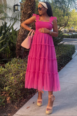 woman wearing a pink tiered ruffle maternity dress, holding her baby bump, accessorized with a pink handbag and sunglasses
