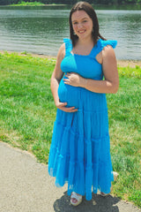 woman wearing a bright blue maternity maxi dress with ruffled straps, holding her baby bump by a lake